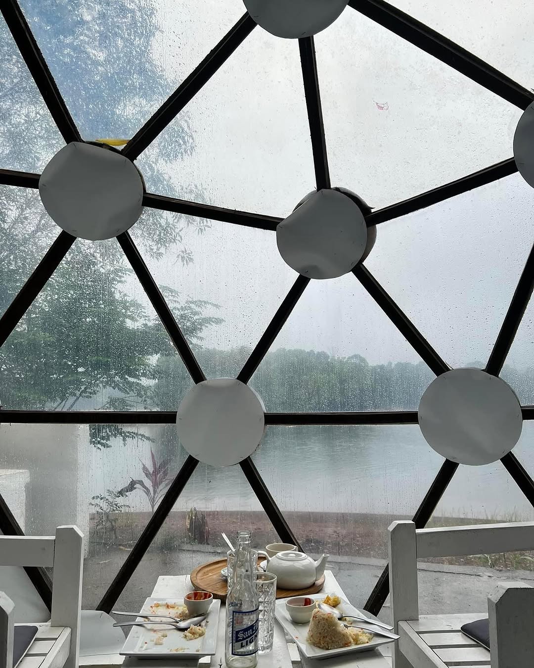 Dome interior — breakfast on table, rain visible through transparent geodesic panels over the lake