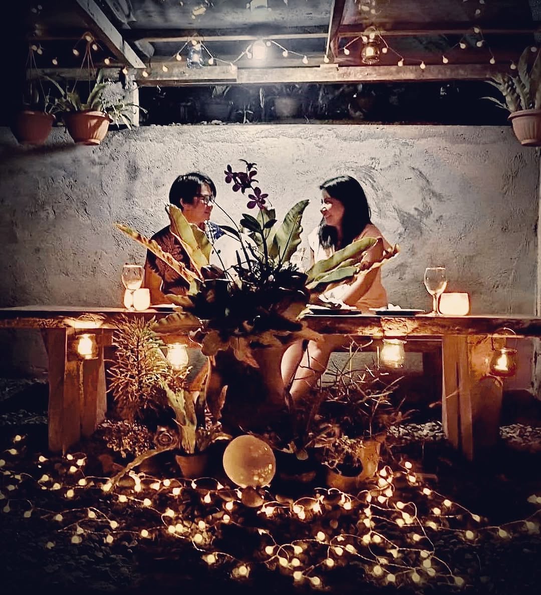 Couple at intimate candlelit dinner table in rustic setting surrounded by fairy lights and plants